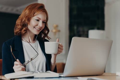 intelligent female student has online course, concentrated in screen of laptop computer, learns foreign languages distantly, drinks tea or coffee, poses at desktop. woman worker has video conference