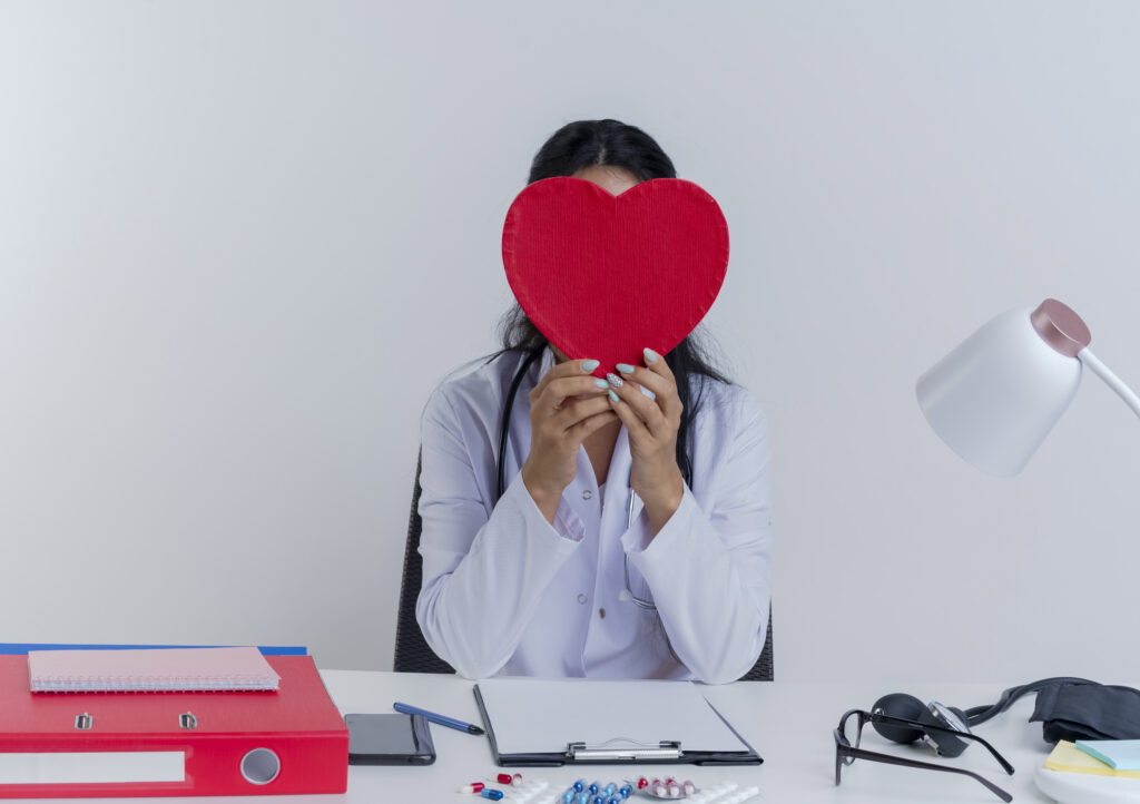 La Neurobiología del Amor y la Amistad: Un Enfoque desde la Neuropsicología young female doctor wearing medical robe and stethoscope sitting at desk with medical tools holding and hiding behind heart shape isolated on white background